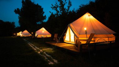 Tents with burning torches, lamps and wooden chairs at glamping, forest around, night