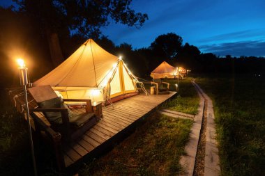 Tents with burning torches, lamps and wooden chairs at glamping, forest around, night