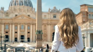 View of the blonde woman from behind in Vatican City. Saint Peter Square with multiple tourists, Saint Peter Basilica and the Vatican Obelisk on the background. Slow motion