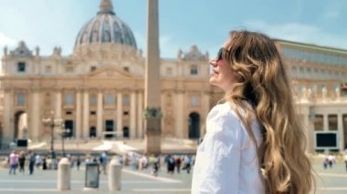 View of the blonde woman from behind in Vatican City. Saint Peter Square with multiple tourists, Saint Peter Basilica and the Vatican Obelisk on the background. Slow motion