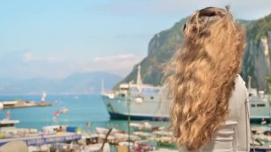 Capri, İtalya 'da poz veren bir kadın. Arka planda tekne ve feribot olan liman manzarası. Yavaş çekim