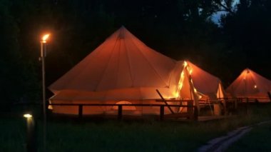 Tents with burning torches, lamps and wooden chairs at glamping, forest around, night