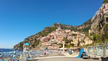 POSITANO, İtalya - Haziran 2022: Tyrhenian deniz kıyısı manzarası. Sahilde insanlar, güneşlikler, tepelerde evler.