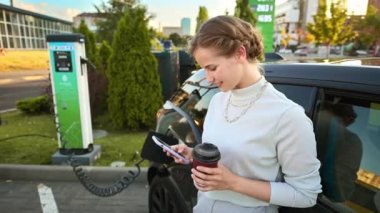 A young blonde woman with smartphone and coffee at a car charging station with charging electric car nearby in Chisinau, Moldova