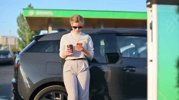 Young Blonde Woman Smartphone Coffee Car Charging Station Electric Car — Vídeos de Stock