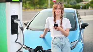 A young blonde woman using smartphone at a car charging station with charging electric car nearby. Chisinau, Moldova. Slow motion
