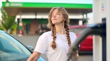 A young blonde woman using smartphone at a car charging station with electric car nearby. Chisinau, Moldova. Slow motion