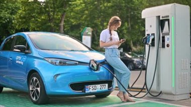 CHISINAU, MOLDOVA - JULY, 2022: A young blonde woman using smartphone at a car charging station with charging electric Renault ZOE. Slow motion