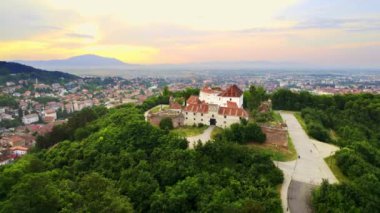 Aerial drone view of The Citadel in Brasov at sunset, Romania. Medieval fortress on the top of a hill. Buildings and green trees around