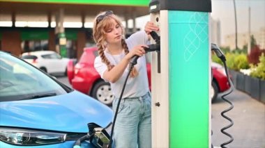 CHISINAU, MOLDOVA - JULY, 2022: A young blonde woman putting a charger into a car charging station, electric Renault ZOE nearby. Slow motion