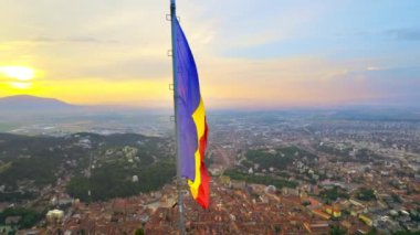 National flag located on the top of the hill with green trees. View of Brasov with multiple buildings on the background at sunset, Romania