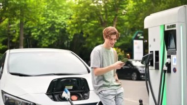 A young man using smartphone at a car charging station with charging electric car nearby. Chisinau, Moldova. Slow motion