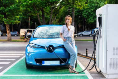 A young blonde woman posing at a car charging station with electric car nearby. Chisinau, Moldova