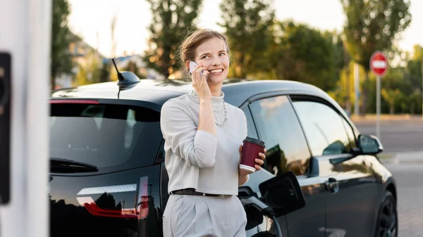 A young blonde woman with coffee talking on the smartphone at a car charging station with charging electric car nearby in Chisinau, Moldova