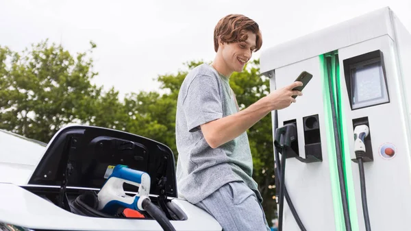 A young man with smartphone at a car charging station and charging electric car nearby. Chisinau, Moldova