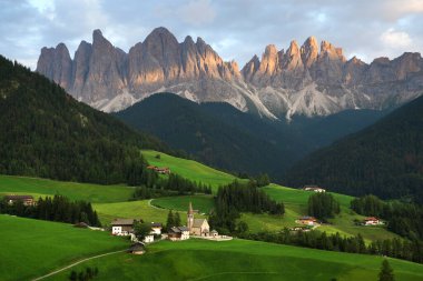 Dolomite Alps at sunset in Italy. Valley with a lot of greenery and houses, slopes covered with forest, view of mountains on the background