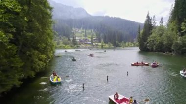 RED LAKE, ROMANIA - AUGUST, 2022: Aerial drone view of nature. Lake in a valley in Carpathian mountains, multiple people floating on boats, lush forest around, village in the distance