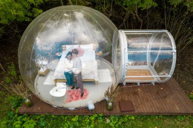 Glamping 'de şeffaf kabarcık çadırı, etrafındaki ve içindeki yemyeşil orman. Birbirine sarılan bir çift.