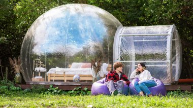 Sandalye çantasına oturmuş glamping 'de sohbet eden bir çift. Arka planda şeffaf kabarcık çadırı, çevrede yemyeşil orman.