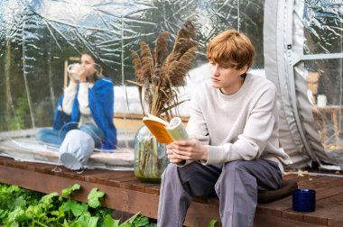 Glamping 'de şeffaf bir kabarcık çadırının yanında kitap okuyan bir adam. Kadın battaniyenin altında, arka planda fincan var.