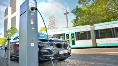 FRANKFURT, ALMANY - Eylül 2022: BMW X5 'i elektrikli araba şarj istasyonunda şarj etmek, arka plandaki tramvayı hareket ettirmek