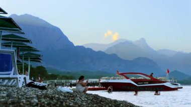 KEMER, TURKEY - 2022 EYLÜL: günbatımında Akdeniz kıyısı sahili görüntüsü. Dinlenen insanlar, rıhtımlar, tekneler, yeşillikler, dağlar