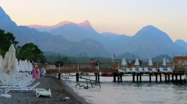 KEMER, TURKEY - 2022 EYLÜL: günbatımında Akdeniz kıyısı sahili görüntüsü. Dinlenen insanlar, iskeleler, yeşillikler, uzaktaki dağlar