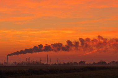 a long tail of smoke streaming across the sky from a refinery against a red sky during sunset. pollution of the atmosphere and the environment by chemical plants, technological development having a negative impact on the environment