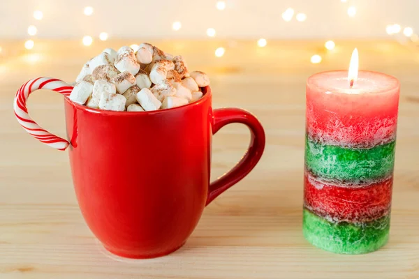 Heiße Schokolade im Becher mit Marshmallows und Zuckerrohr, Weihnachtskerze auf dem Tisch mit Weihnachtsbeleuchtung. — Stockfoto