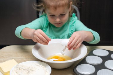 Mutfakta yemek yapan küçük bir kız. Çocuk elinde yemekle masada oturuyor. Çocuk evde yemek pişiriyor, kek yapmayı öğreniyor, kasede yumurta kırıyor..