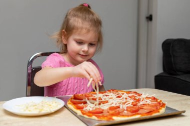 Evde yemek yapan küçük bir çocuk masada oturuyor. Küçük kız pizza yapıyor, peynir ekliyor. Çocuk yemek yapmayı öğreniyor.