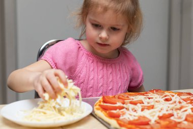 Small child cooking meal at home sitting at the table in the kitchen at home. Little girl making pizza, adding cheese.