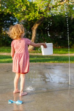 Küçük kız yazın su sıçratma yerinde su ve oyuncaklarla oynuyor. Güneşli bir günde fıskiyeli çocuk bahçesinde.