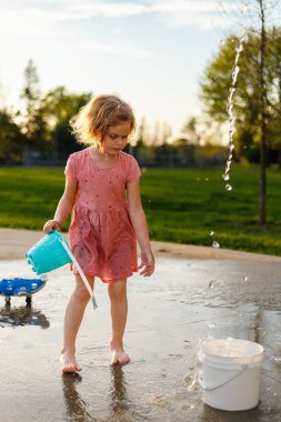 Küçük çocuk yazın fıskiyelerle parkta su ve oyuncaklarla oynuyor. Küçük çocuk su sıçratma parkında eğleniyor.