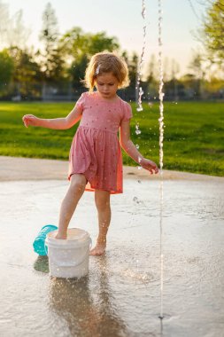 Küçük kız yazın fıskiyelerle parkta oynuyor. Küçük çocuk su sıçratma alanında eğleniyor.