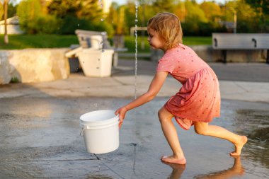Çocuk su sıçratma yerindeki parkta kova ve suyla oynuyor. Kız yazın fıskiyelerle eğleniyor.
