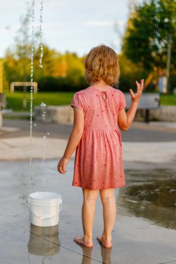 Küçük kız yazın su sıçratma yerinde suyla oynuyor. Çocuk parkta fıskiyeli çocuk