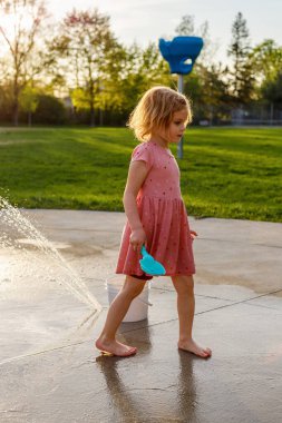 Küçük kız yazın parkta su sıçratma yerindeki çocuk parkında oyuncaklarıyla oynuyor. Çeşmelerin yanındaki küçük çocuk dışarıda vakit geçiriyor.