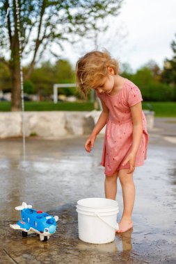 Küçük kız yazın parkta su sıçratma yerindeki çocuk parkında oyuncaklarıyla oynuyor. Çeşmelerin yanındaki küçük çocuk dışarıda vakit geçiriyor.