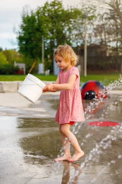 Child playing with bucket at splash pad playground in water park. Girl with fountain in summer.