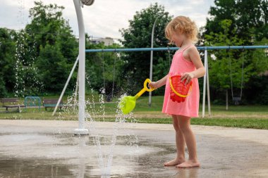 Küçük çocuk sıcak yaz günü yerel halk parkında su ve oyuncaklarla oynuyor. Pembe elbiseli küçük güzel kız çeşme parkında eğleniyor.