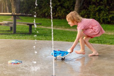 Küçük kız yazın parkta su sıçratma yerindeki çocuk parkında oyuncaklarıyla oynuyor. Çeşmelerin yanındaki küçük çocuk güneşli bir günde dışarıda vakit geçiriyor.