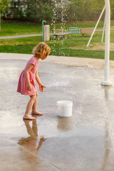 Child playing with water and bucket at splash pad playground in park. Girl with fountain in summer.