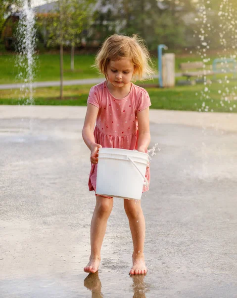 Child playing with water and bucket at splash pad playground in park. Girl with fountain in summer.