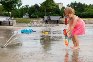Sıcak bir yaz gününde, şehir parkında su ve oyuncaklarla oynayan küçük bir çocuk. Pembe elbiseli küçük güzel kız çeşme parkında eğleniyor.