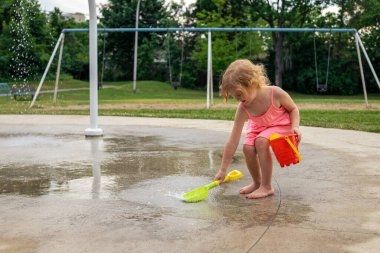 Sıcak bir yaz gününde, şehir parkında su ve oyuncaklarla oynayan küçük bir çocuk. Pembe elbiseli küçük güzel kız çeşme parkında eğleniyor.