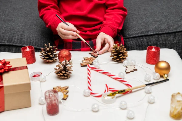 Christmas kids craft activities at home. Little child painting pine cones and wooden Christmas tree decorations at home. Holiday DIY concept for children.