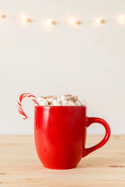 Hot chocolate in red mug with small marshmallows on wooden table with holiday lights on wall. Cocoa in a cup, hot winter drink