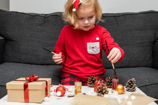 Christmas kids craft activities at home. Little child painting pine cones and wooden Christmas tree decorations at home. Holiday DIY concept for children.