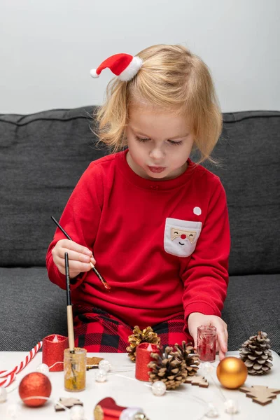 Christmas kids craft activities at home. Little child painting pine cones and wooden Christmas tree decorations at home. Holiday DIY concept for children.